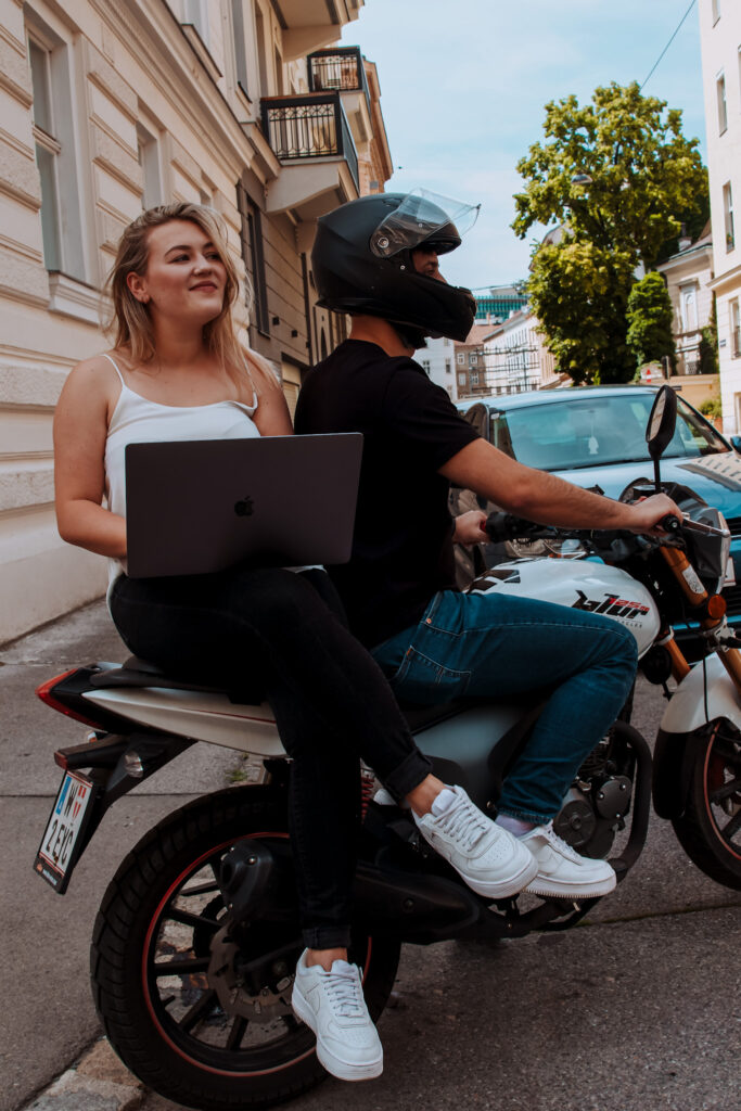 Iris und Kaan auf dem Motorrad. Iris sitzt hinten und hat einen Laptop in der Hand.